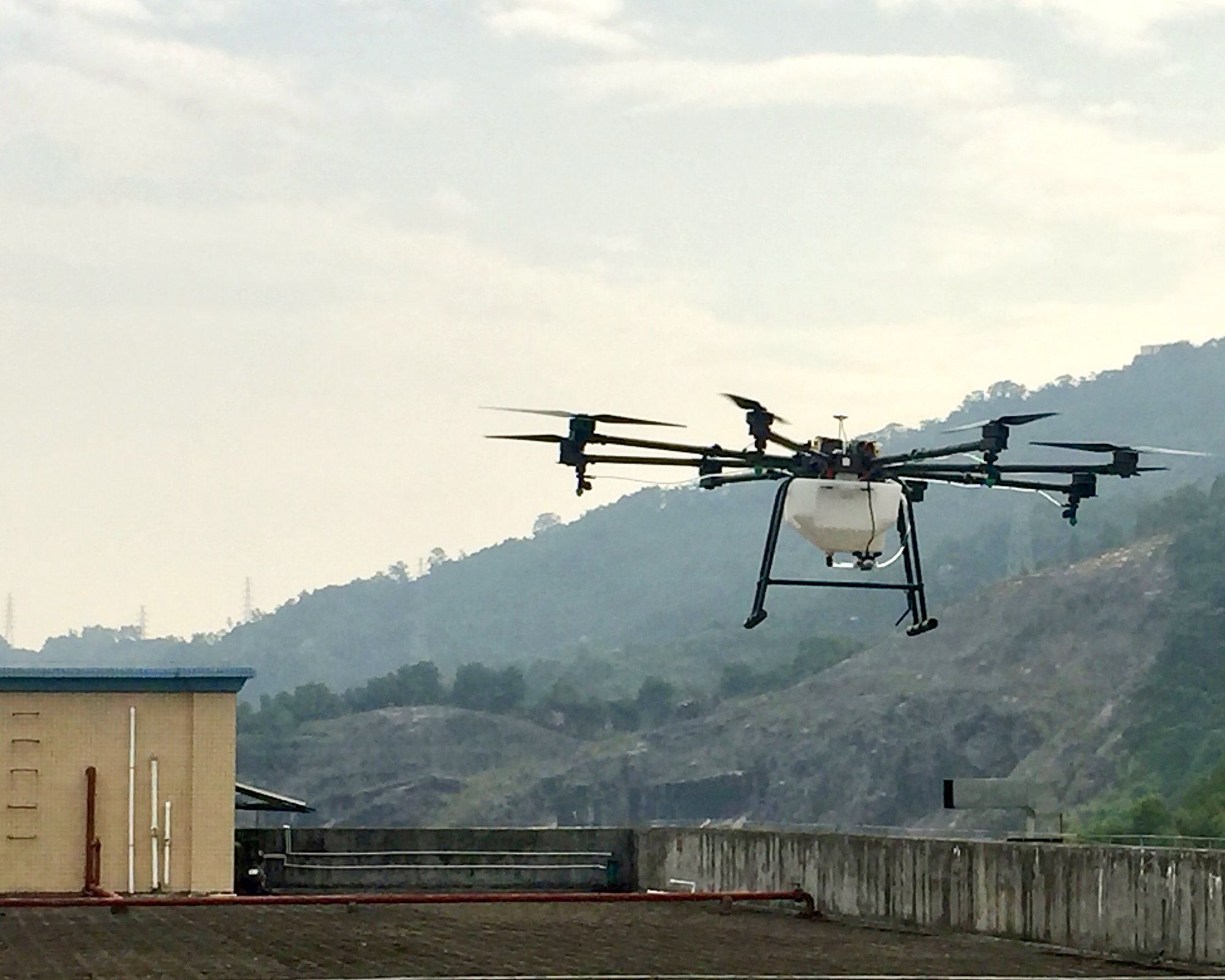 Eight-rotor agricultural spray drone with a white tank in a test flight over a factory roof